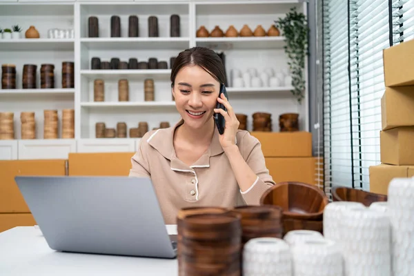 Asian beautiful woman talk on smartphone to sell vase product at home. Young attractive girl use laptop computer shows goods to customer and present detail. Remote buying and purchase shopping concept