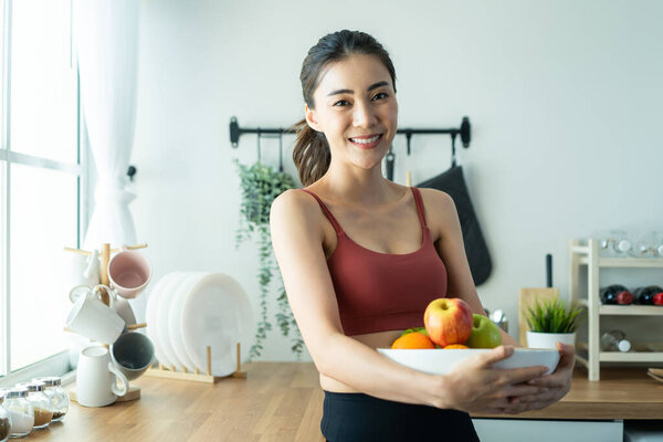 Asian attractive active woman holding bowl of fruit in kitchen at home. Portrait of active sport girl in sportswear enjoy eat apple healthy food for health after exercise and look at camera in house.