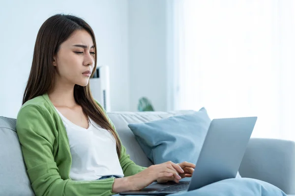 Exhausted Asian business woman hurt eye while using laptop computer. Young girl office worker sit on sofa feel visual fatigue and eye strain tired from overwork and massage dry irritable eye at home.