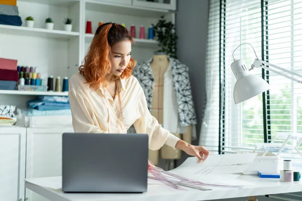 Asian tailor woman working to design new clothes in tailoring atelier. Attractive young female fashion designer dressmaker create new pieces of clothing from patterns and designs in workshop room.