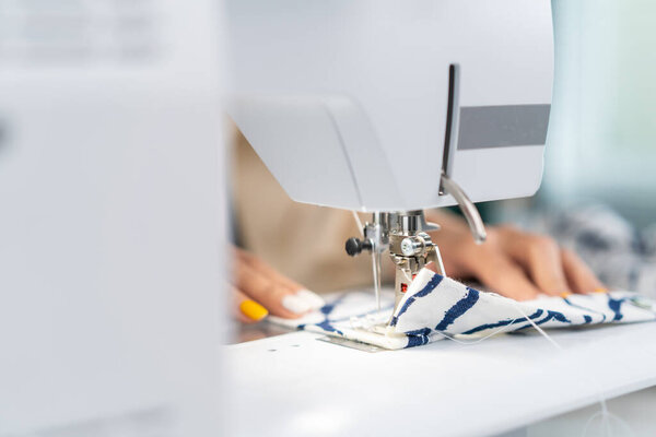 Close up hands of designer pushes fabric material in sewing machine. Beautiful young fashion tailor woman working to making a new handmade clothes for collection in workshop room or tailoring atelier.