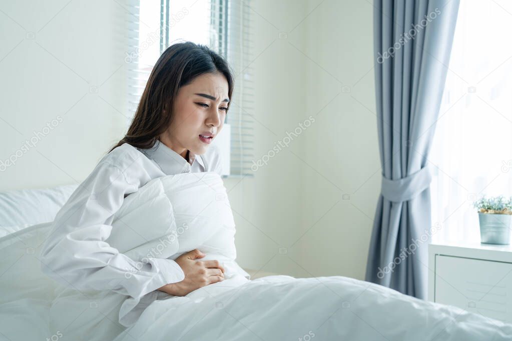 Asian beautiful sick girl in pajamas getting up from sleep in bedroom. Attractive young woman feel bad and painful after wake up on bed, having stomach ache or periods then clasping belly in morning.