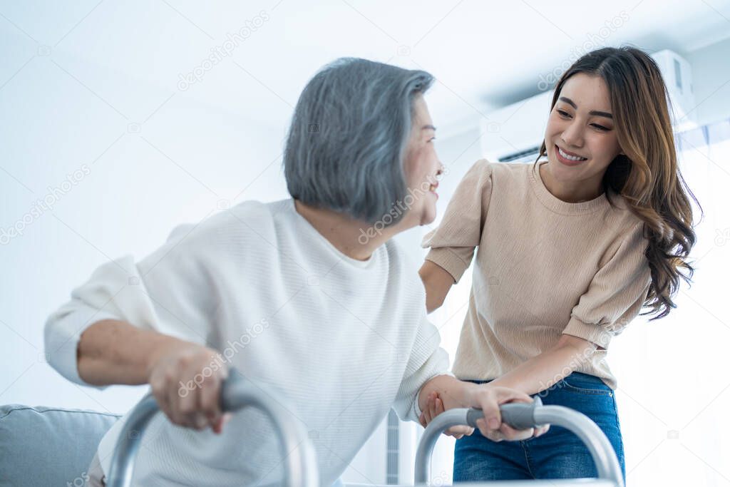 Asian young daughter support old disabled woman walk with walker at home. Beautiful girl help and take care of senior elderly mature handicap mom patient doing physical therapy in living room in house