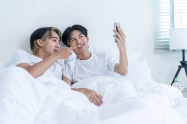 Asian handsome man gay use mobile phone video call online with family. Attractive handsome LGBTQ in pajamas lying down on bed in bedroom, hold smartphone and talk virtual remote in morning in house.