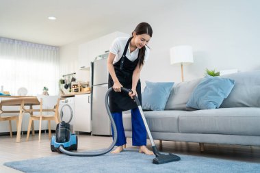 Asian cleaning service woman worker cleaning in living room at home. Beautiful young girl housekeeper cleaner wear apron and vacuuming messy dirty floor for housekeeping housework and chores in house.