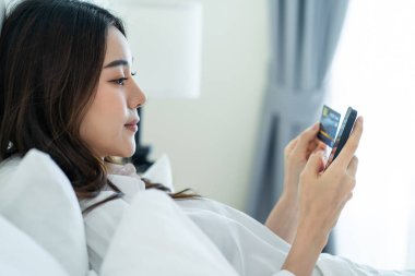 Asyalı çekici kadın sabah evde cep telefonu sohbetini kullanıyor. Mutlu, sıradan, güzel bir kadın yatak odasında uzanıyor. Tatil haftasonunun tadını çıkarın. Evdeki teknolojiyle akıllı telefon iletişimi kurun..