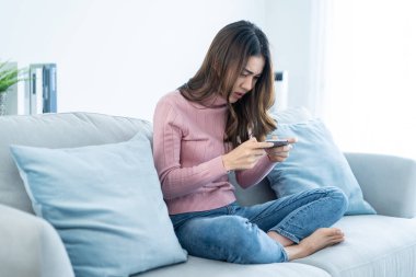 Asian frustrated beautiful woman play mobile game on cellphone at home. Attractive casual e-sport gamer girl loser sit on couch in living room, feel angry and offended due to defeat in the video game.