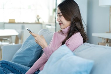 Asyalı güzel kadın kanepede oturur ve cep telefonuyla sohbet eder. Mutlu çekici genç kız evde vakit geçirir, rahatlar, iletişim kurar ve oturma odasında akıllı telefondan tartışır..