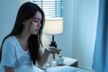 Asian unhappy sick girl in pajamas take medicine before sleep on bed. Attractive beautiful young woman feel frustrated to take many drug to in dark night in bedroom at home. Healthcare-medical concept