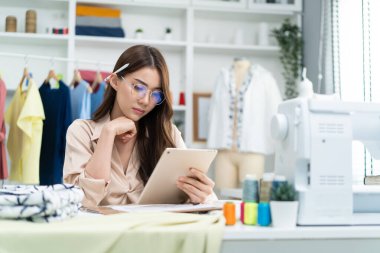 Asian tailor woman working to design new clothes in tailoring atelier. Attractive young female fashion designer dressmaker use tablet to find reference to think and design new clothes in workshop room
