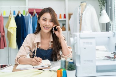 Asian tailor woman talking on phone with customer in tailoring atelier. Attractive beautiful young female fashion designer working and checking new fashionable handmade clothes collection in workshop.