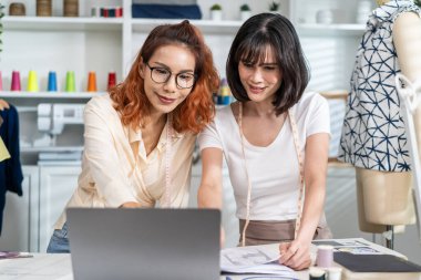 Asian two tailor woman working to design clothes in tailoring atelier. Attractive young female fashion designer dressmaker using laptop computer work to create new pattern collection in workshop room.
