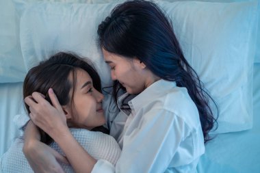 Asian beautiful lesbian couple lying down on bed and hugging each other. Attractive romantic girl friend in pajamas spending night leisure time together in bedroom. Homosexual freedom pride concept.