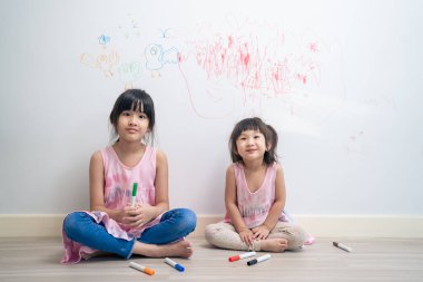 Asian young sibling kid girl enjoy paint on white wall in living room. Little adorable children having fun drawing and coloring art picture with hapiness enjoy creativity at home and looking at camera