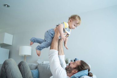 Caucasian loving dad play with baby boy child on sofa in living room. Happy family, attractive handsome caring young father lifting toddler son fly up enjoy doing airplane game in morning in house.
