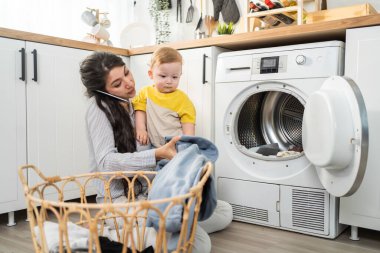 Beyaz tenli anne mutfakta bebek bebekle ev işi yapıyor. Güzel anne iş için telefon eder ve oğluyla evde oynaşmak için çamaşır makinesine çamaşır koyar. Aile Hizmetleri.