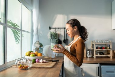 Latin çekici kadın evdeki mutfakta önlük aşçısı yeşil salata giyiyor. Genç ve güzel bir kız kendini mutlu hissediyor ve sebze yemekten hoşlanıyor. Diyet için sağlıklı yiyecekler ve evde sağlık hizmetleri için kilo vermek..