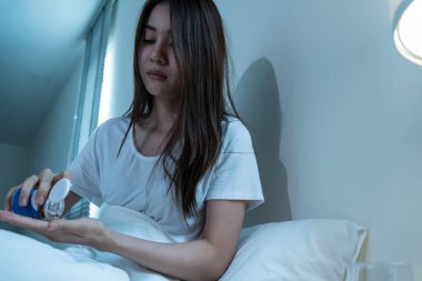 Asian unhappy sick girl in pajamas take medicine before sleep on bed. Attractive beautiful young woman feel frustrated to take many drug to in dark night in bedroom at home. Healthcare-medical concept
