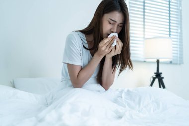 Asian sick girl in pajamas wake up from sleep at night sneezing on bed. Attractive young woman feeling bad and suffer from allergy, put tissue cover her nose while sneezes during bedtime in house.