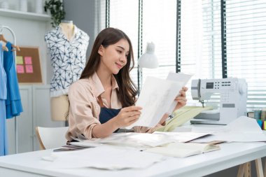 Asian beautiful tailor woman working on clothes in tailoring atelier. Attractive female fashion designer dressmaker design drawing and sketching out ideas for new fashion collection in workshop room.