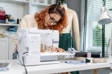 Asian beautiful tailor woman working on clothes in tailoring atelier. Attractive young female fashion designer pushes fabric material through a sewing machine making new handmade suit in workshop room