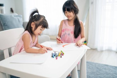 Asian young girl children drawing and coloring paper in living room. Little adorable sibling child sister sit on table, learn how to sketch art picture enjoy creativity activity on holiday at home.