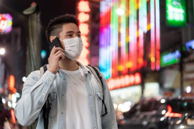 Asian attractive man backpacker talking on mobile phone walk in city. Young male tourist traveler wear mask travel alone on street, using smartphone enjoy spend time on holiday vacation trip at night.