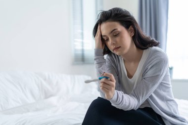 Asian young woman feeling shock after look at positive Pregnancy test. Disappointed attractive beautiful unplanned pregnancy girl sit on bed with upset and sad face for problem don't want to pregnant.