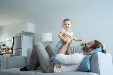 Caucasian loving dad play with baby boy child on sofa in living room. Happy family, attractive handsome caring young father lifting toddler son fly up enjoy doing airplane game in morning in house.