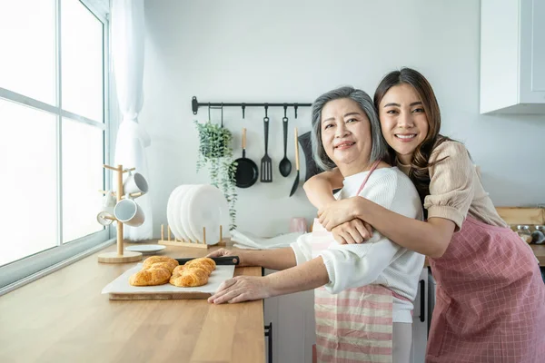 Portrait of Asian lovely family, young daughter stand with old mother. Attractive female and senior elderly mom wear apron enjoy activity at home to bake croissant in kitchen and smile, look at camera