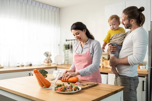 Caucasian beautiful parents cook food with baby boy toddler in kitchen. Happy family, Attractive young mother making healthy salad for lunch while husband father holding little kid son infant in house