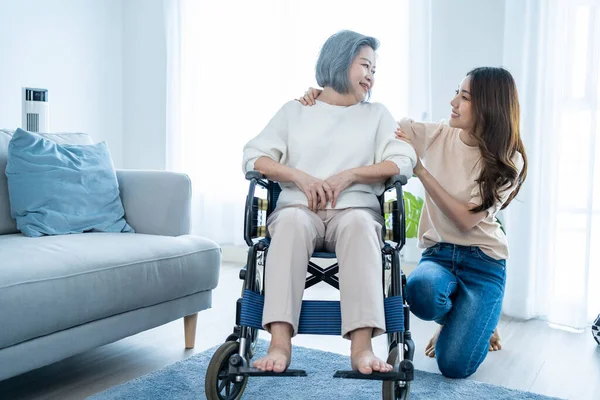 Asian young daughter support older senior woman on wheelchair at home. Beautiful girl granddaughter help and take care of elderly mature mother patient doing physical therapy in living room in house.
