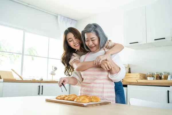 Asian lovely family, young daughter look to old mother cook in kitchen. Beautiful female enjoy spend leisure time and hugging senior elderly mom bake croissant on table in house. Activity relationship