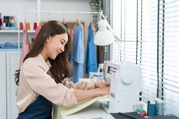 Asian beautiful tailor woman working on clothes in tailoring atelier. Attractive young female fashion designer pushes fabric material through a sewing machine making new handmade suit in workshop roomc
