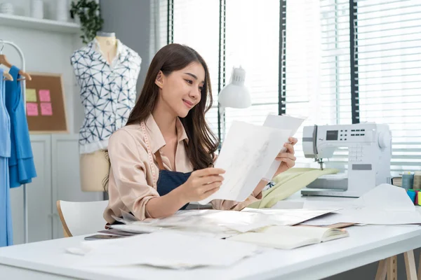 Asian beautiful tailor woman working on clothes in tailoring atelier. Attractive female fashion designer dressmaker design drawing and sketching out ideas for new fashion collection in workshop room.