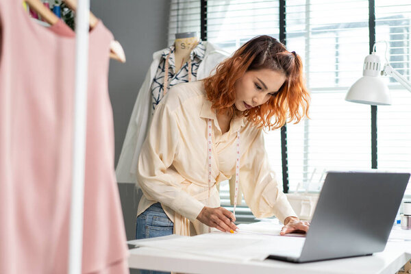 Asian beautiful tailor woman working on clothes in tailoring atelier. Attractive female fashion designer dressmaker design drawing and sketching out ideas for new fashion collection in workshop room.