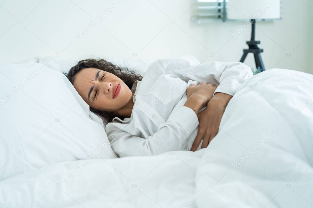 Latino beautiful sick girl in pajamas getting up from sleep in bedroom. Attractive young woman feel bad and painful after wake up on bed, having stomach ache or periods then clasping belly in morning.