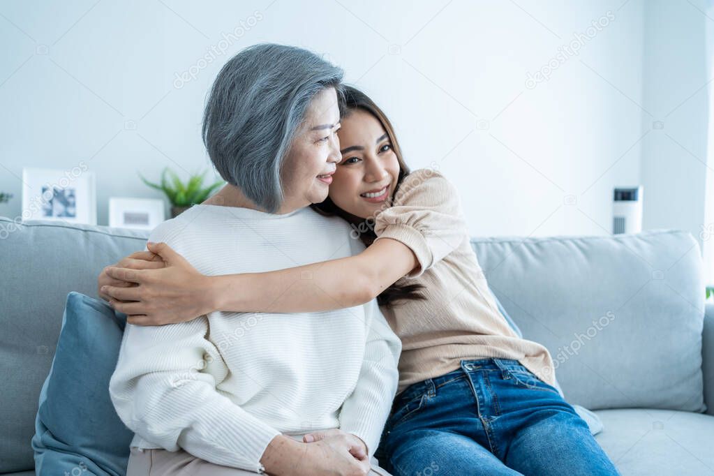 Portrait of Asian lovely family, young daughter hugging older mother. Attractive beautiful woman and senior elder mature mom sitting on sofa with smile, enjoy activity at home together in living room.