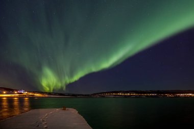 Drapery Aurora - Northern lights over a fjord with a snow covered pier in the foreground and a illuminated church in the background in Northern Norway