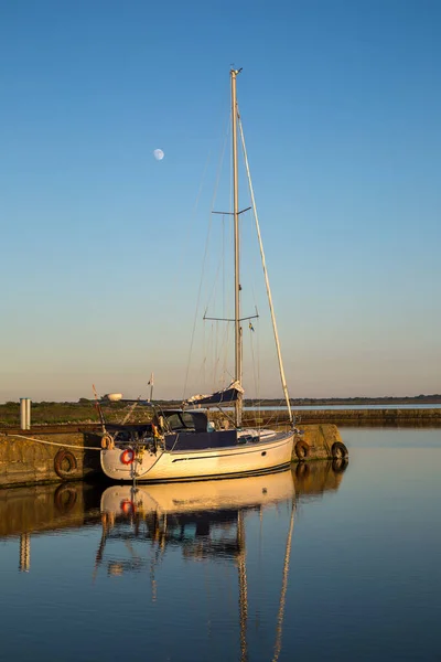 Günbatımında Gronhogen marinasındaki rıhtıma demirleyen yelkenli, sakin suda yansıyor ve ay berrak bir gökyüzünde yükseliyor. Oland, İsveç.