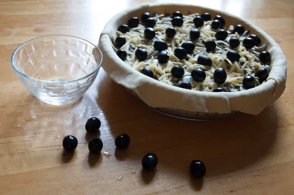 Préparation de tarte (la pâte, les olives et le fromage râpé dans un plat de cuisson en verre, près d'une tasse en verre, quelques olives sur la table ) — Photo