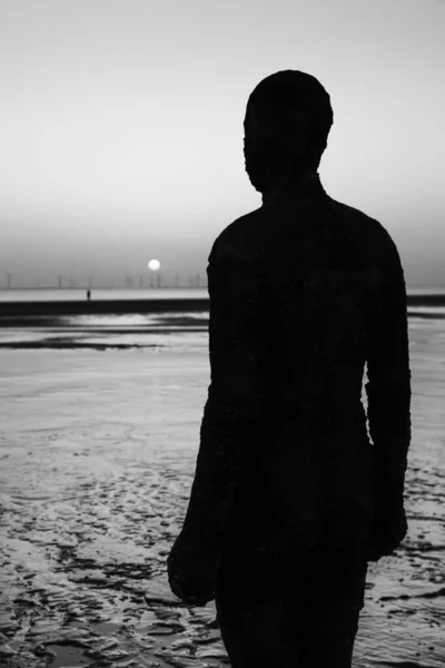 Monochrome silhouette of an Iron Man statue (one of the one hundred Iron Men statues which make up Antony Gormley\'s Another Place art installation) captured on Crosby Beach near Liverpool at sunset in April 2021.