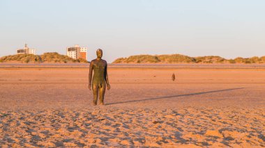 Nisan 2021 'de Crosby plajında gün batımında Demir Adam' ın arkasında uzun bir gölge. Bu ve arkadaki, Liverpool yakınlarında Antony Gormley tarafından yaratılan 100 Demir Adam heykelinden oluşan başka bir yerin parçası..