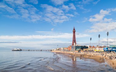 Eylül 2021 'de Blackpool plajında sular yükselmeye başladığında turistler suya giriyor. Kuzey İskelesi ve Blackpool Kulesi mavi gökyüzünün altında yer alır..