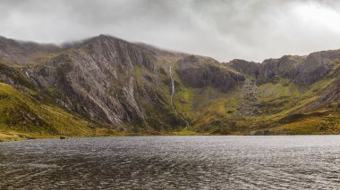 Güneş ışığı, Snowdonia Ulusal Parkı 'ndaki Idwal Gölü' nün üzerindeki kara bulutları delip geçer. Dağ yamacını aydınlatır ve yağmur suyu dereleri aşağı akar..