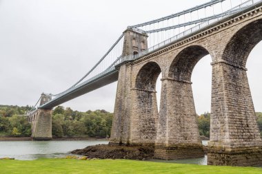 Kuzey Galler ve Anglesey arasındaki Menai Boğazı boyunca uzanan devasa Menai Askı Köprüsü 'ne bakıyorum..