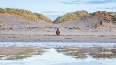 Crosby Sahili 'ndeki kum tepeleri arasında görülen bir Demir Adam. Antony Gormley tarafından kumsalda yaratılan diğer 99 figürle birlikte 2022 yılının Ocak ayının akşam karanlığından önce ıslak kuma yansıdığı görüldü..
