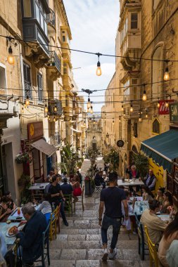 Nisan 2022 'de Valletta' da popüler bir restoranın önünde yemek yiyen turistlerle dolu merdivenlerin üzerinde ışıklar asılı..