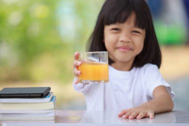 Asyalı Taylandlı kız parlak gülüşlü, beyaz tişörtlü, sağlıklı bir kız. Dışarıda oturuyor. Masanın üzerinde kitaplar ve bir bardak portakal suyu var. Arka plan bulanık yeşil..