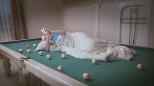 Man with a cell phone in his hands lies on a pool table, covered with a blanket — Stock Video